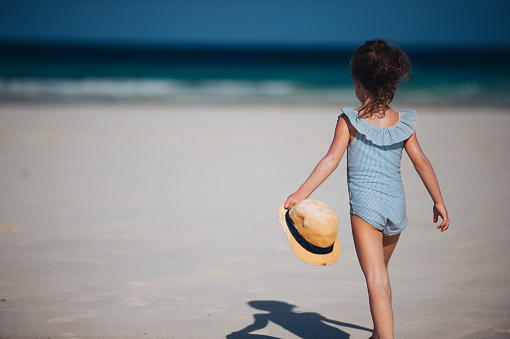Rear view of a little girl walking on the beach in a swimsuit and a straw hat in hand. Full body shot of smilling girl in blue swimsuit, enjoying sandy beach and crystalline sea of Mnemba beach in Zanzibar, with copy space. Concept of beach summer vacation with kids.