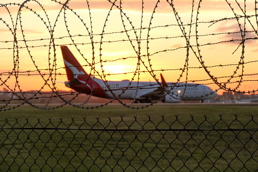 A Qantas Boeing B737-838 plane, registration VH-VYZ, taxiing after landing at Sydney Kingsford-Smith Airport as flight QF541 from Brisbane.  In the foreground is barbed wire, razor wire and mesh wire that forms the airport boundary.  This image was taken from near Shep's Mound, Ross Smith Avenue, Mascot after sunset on 25 August 2023.