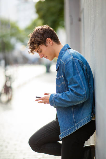 istock Young man using mobile phone leaning against gray wall outside 1637958966
