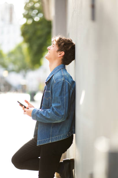 Smiling young man with cell phone leaning against a wall outdoors stock photo