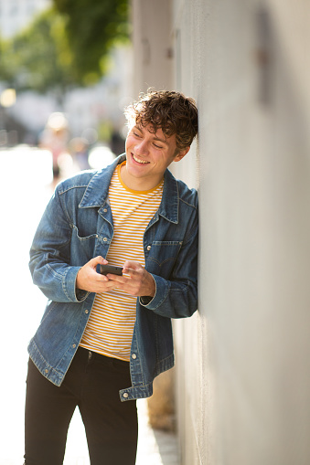 Portrait of stylish young man with mobile phone leaning to a wall in city and smiling