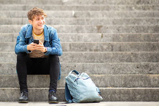 Portrait of handsome young man sitting on steps outside with phone and bag looking away smiling