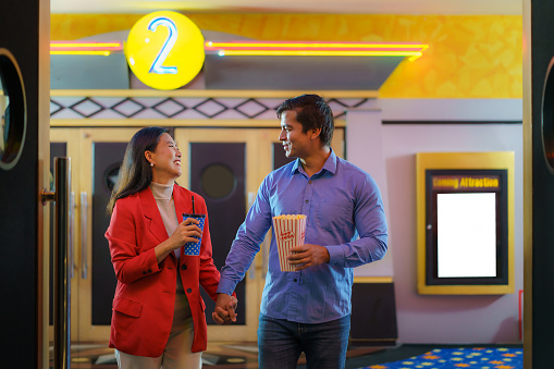 Radiating joy, an interracial couple holding hands walks into the cinema, drinks in hand, ready to savor a movie night with popcorn and togetherness.