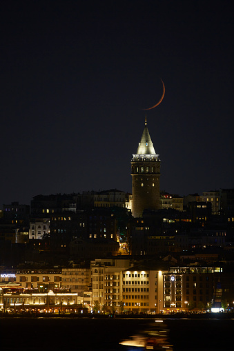 Istanbuls best known place Galata Tower with a moon setting over and passing by ferries in motion and light