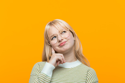 Blond hair teenage girl wearing green sweater and eyeglasses standing with hand on chin and looking up. Studio shot, yellow background.