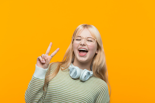 Happy blond hair teenage girl wearing green sweater, headphone and eyeglasses showing peace sign and laughing at camera. Studio shot, yellow background.