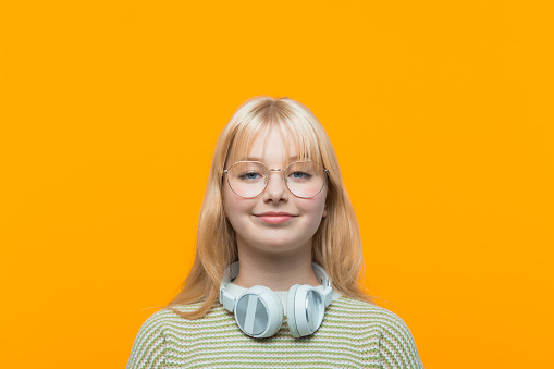 Happy blond hair teenage girl wearing green sweater, headphone and eyeglasses smiling at camera. Studio shot, yellow background.