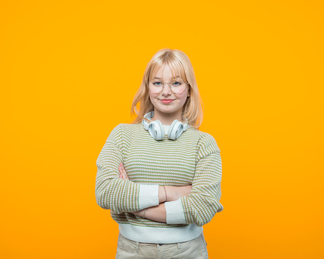 Blond hair teenage girl wearing green sweater, headphone and eyeglasses standing with arms crossed and smiling at camera. Studio shot, yellow background.