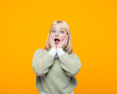 Excited blond hair teenage girl wearing green sweater and eyeglasses standing with face in hands and staring at camera. Studio shot, yellow background.