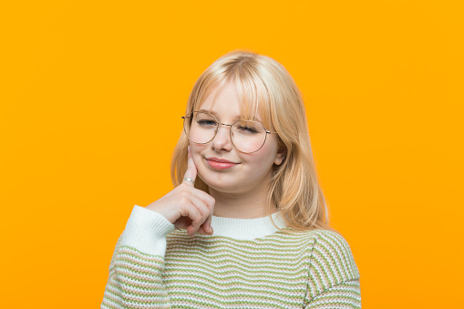 Blond hair teenage girl wearing green sweater and eyeglasses standing with hand on chin and smiling at camera. Studio shot, yellow background.