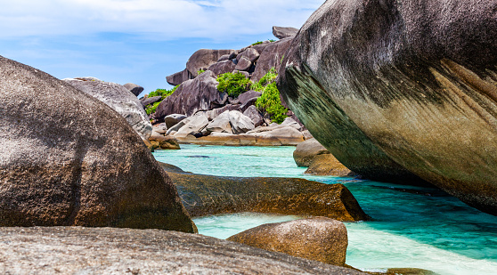 A serene view of a tropical beach of Similan islands with bright blue water and white sand. The water is very clear, you can see the rocks and sand on the bottom. There are scattered gray boulders throughout the image, they are smooth and round. The sky is a light blue with some white clouds. The image conveys a sense of peace and serenity.