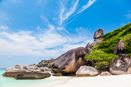 A scenic view of a tropical beach on Similan islands with white sand and clear blue water. On the right side of the image, there are large dark grey boulders covered with dense green vegetation. The sky is blue with white clouds. The image is taken from a low angle, looking up at the boulders and greenery.