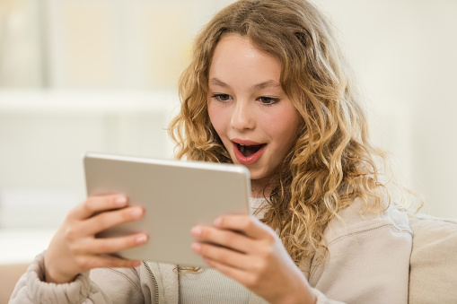 Side view of surprised teenage girl sitting on sofa in the living room at home, holding digital tablet in hands.