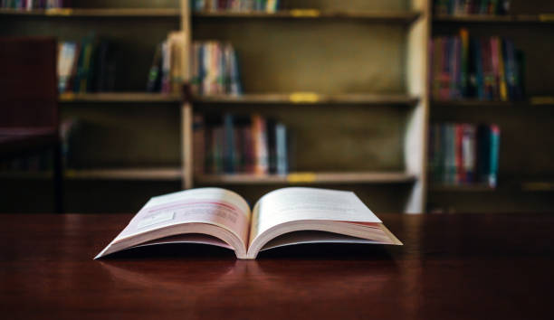 istock On a wooden table, there's an open book with a library visible in the background. A stack of books nearby adds to the theme of educational learning. 1638106548