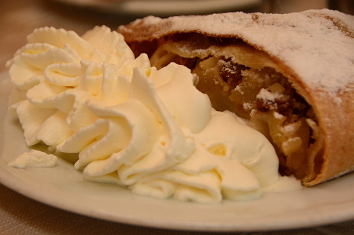 Pastry: Apple strudel behind a heap of whupped cream, served on a plate. Layered dough pastry with a apple filling inside.\nPiece of a pie.