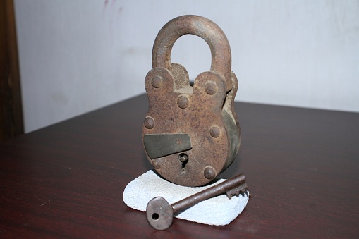 An old designed padlock on a dark coloured table with white background, the padlock has a key also, the padlock stands on a piece of small stone with the key, the padlock and the key are made of metal and it is very old so it is rusty.