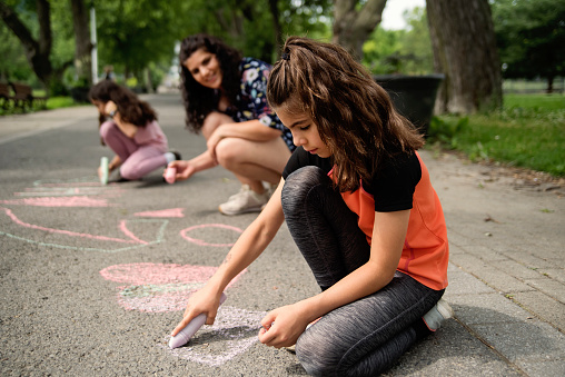 Mother of middle-eastern ethnicity playing with two active daughters in park. She is in her early forties, girls are 8 and 9 year’s old. They are drawing on the pavement. Horizontal full length outdoors shot with copy space. This was taken in Montreal, Quebec, Canada.