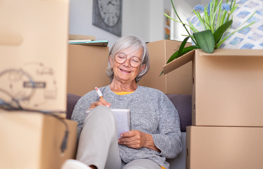 Happy senior woman involved in moving house sitting among cardboard boxes making note of purchases and to-dos, concept of moving, retirement, new life, buying, renting, apartment, house