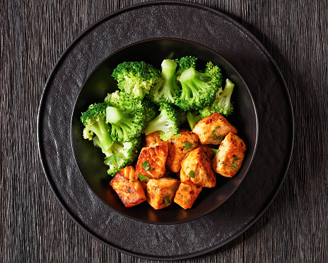 baked salmon bites with steamed broccoli florets in black bowl on dark wooden table with sriracha mayo sauce, horizontal view from above, close-up