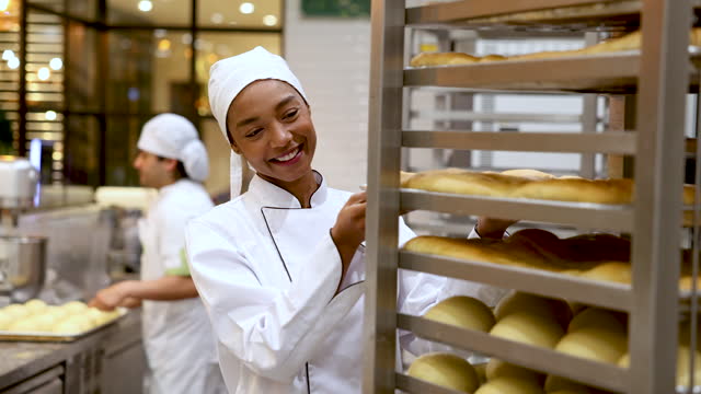 Beautiful black female baker putting a fresh baked tray of bread on moving shelf and then smiles at camera