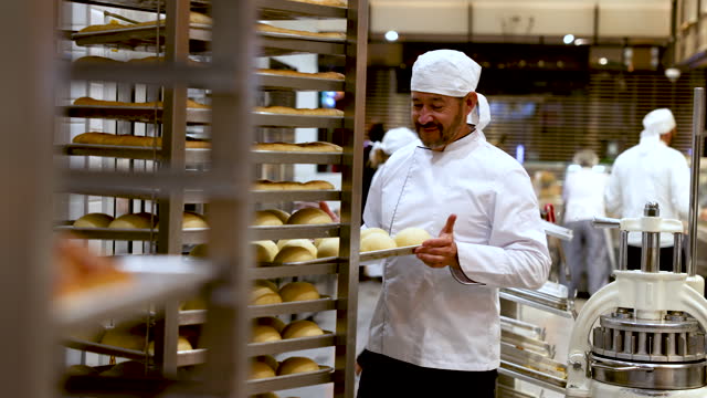 Male baker adding a tray of raw bread on the moving shelf while the rest of the team work at background