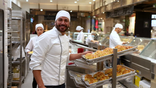 Video portrait of a happy Latin American baker moving trays of fresh baked croissants and then facing the camera smiling
