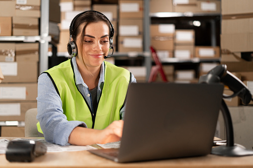 Portrait of a smiling warehouse staff listening and talking to a customer using a headset while working in a distribution warehouse. female call center or support operator working in online stores.