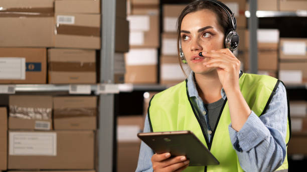 istock brazilian female worker in headset at logistics warehouse using digital tablet. speaks into a microphone against the background of shelves with cardboard boxes. 1638276612