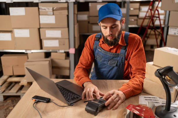 istock Postal warehouse worker using pos-terminal to pay for receiving a parcel or goods sitting at a table with a laptop 1638287406