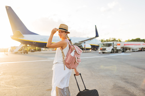 Portrait of a smiling woman in a straw hat walking towards the plane dragging a suitcase, but turned and looked directly at the camera