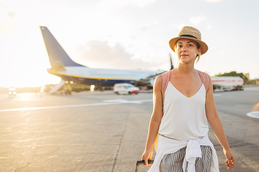 Waist up view of a woman in a straw hat getting off an airplane and dragging a suitcase behind her