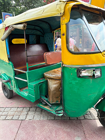 Delhi, India - July 26, 2023: Stock photo showing close-up view stationary, off duty, green and yellow auto rickshaw parking on paving stone footpath surrounded waiting for a passenger fare.