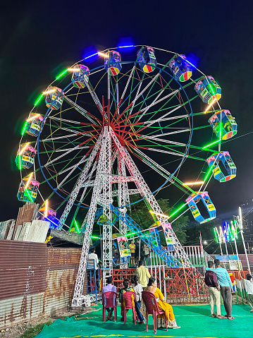 Noida, India - August 13, 2023: Stock photo showing close-up view of Ferris wheel amusement park ride at night illuminated by neon lights with people in line.