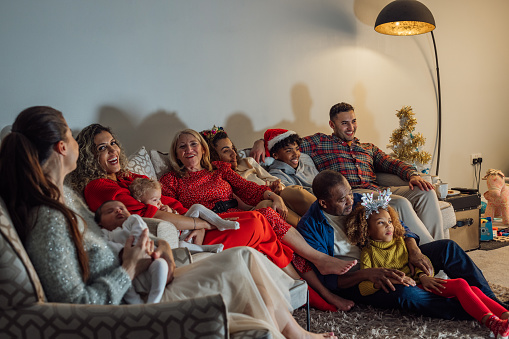 A medium wide angle side view of a family relaxing in their living room sitting watching Christmas TV. They are enjoying time together as a family. They are a multi gen family with three generations in the family home.