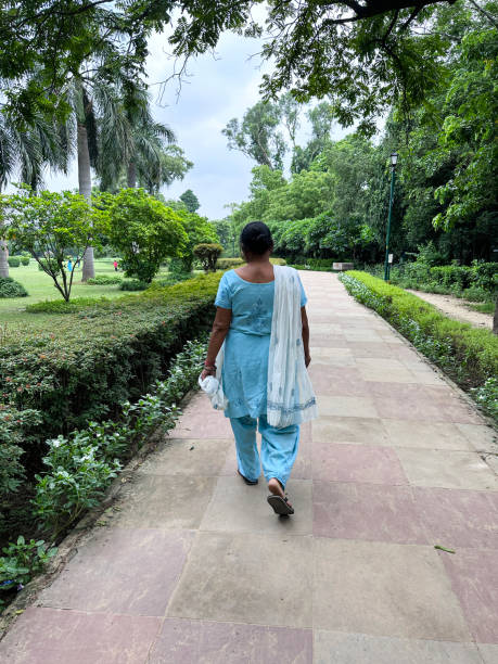 Stock photo showing the free to enter landscaped public park of Lodhi Gardens home to the mausoleums of Mohammed Shah and Sikander Lodhi. imagen de una mujer india vestida con ropa tradicional azul salwar kameez, dupatta blanca (bufanda) sobre los hombros, caminando por un camino pavimentado bordeado de árboles, parque público lodhi gardens, césped verde, macizos de flores cultivados, set