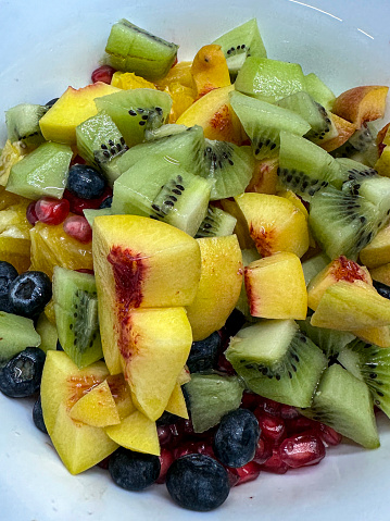 Stock photo showing an elevated view of fresh fruit salad of nectarine, kiwi, pomegranate seeds and blueberries in a white bowl.