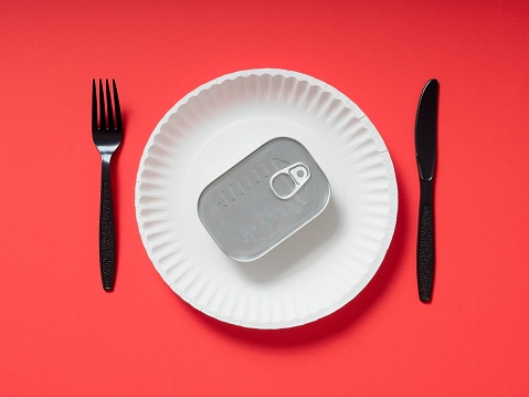 Unopened sardine can on paper plate with plastic ware on red. Design element of eating basic food out of a can with copy space.
