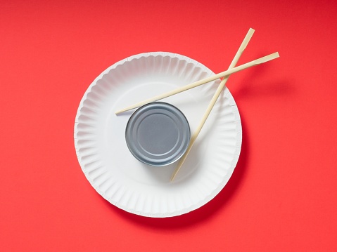 Tuna can on paper plate being picked up by chopsticks on red background. Design element of eating basic food out of a can with copy space.