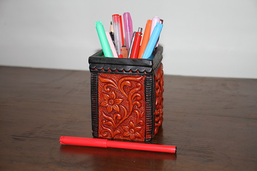 A pen holder on the wooden surface with a white wall, the pen holder holds many coloured pens and pencil, the holder is made of clay painted with colours, the pen holder has a carved design on it, which makes the holder more beautiful.
