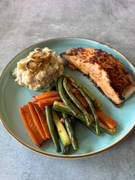 Stock photo showing close-up, elevated view of a healthy meal consisting of a dry fried salmon fillet served with mashed potatoes and roasted vegetables including courgette, carrot and green beans. This dish is part of a healthy eating, low calorie diet plan. imagen de plato moteado de filete de salmón frito a la plancha, puré de papas, guarnición de rodajas de limón, verduras asadas, rodajas de calabacín, porras de zanahoria, judías verdes, fondo gris, dieta de comida saludable, vista elevada, enfoque en