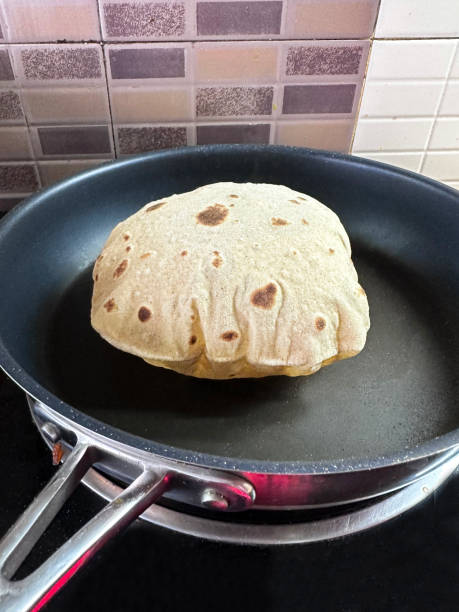 Image of non-stick frying pan containing inflated roti, chapatti made with wholewheat atta flour, cooking on kitchen stove top, tiled wall splash back, elevated view, focus on foreground stock photo