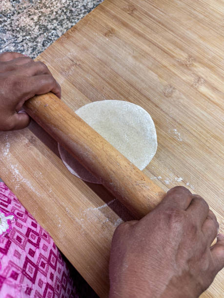 Close-up image of unrecognisable Indian woman preparing a roti with rolling pin on wooden chopping board, kitchen counter, wholewheat atta flour chapatti dough rolled into disk, elevated view stock photo