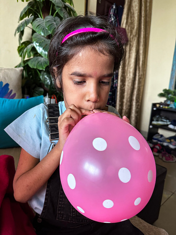 Stock photo showing close-up view of an Indian girl sitting on sofa whilst inflating a party balloon.
