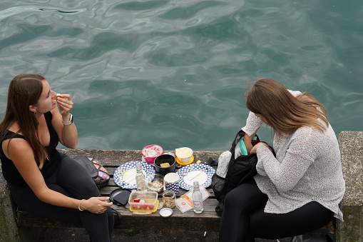 Zurich, Switzerland, 07 02 2023, Two young women making a picnic on the side of Lake Zurich. They sit opposite each other on a cement pier. Between them there are plates, bottles and food containers.