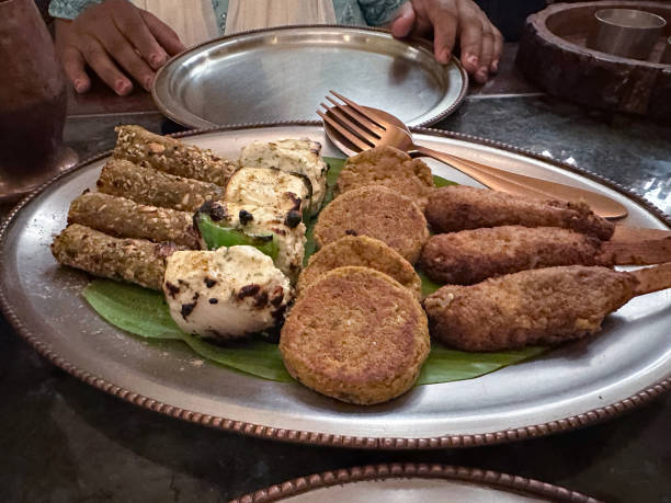 Stock photo showing close-up, view of table set with an appetizer meal served on a metal platter. imagen en primer plano de la mesa con un plato de metal para compartir de bocadillos indios que incluyen paneer, pakoras y kebabs, cuchara y tenedor, platos de metal, mujer india irreconocible vestida con ropa tradicional azul salwar kameez, dupatta blanca