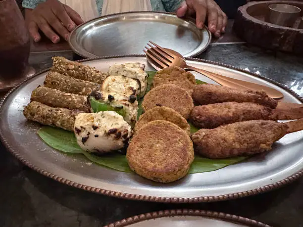 Photo of Close-up image of table set with metal sharing platter of Indian snacks including paneer, pakoras and kebabs, serving spoon and fork, metal plates, unrecognisable Indian woman wearing blue Salwar Kameez traditional clothing, white dupatta (scarf)