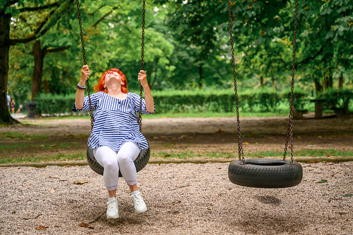Cheerful senior woman with long red hair enjoying and having fun on a swing in a green public park on a summer day.