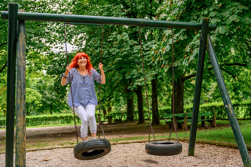 Cheerful senior woman with long red hair enjoying and having fun on a swing in a green public park on a summer day.