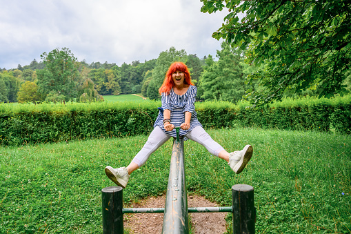 Cheerful senior woman with long red hair enjoying and having fun on a swing in a green public park on a summer day.