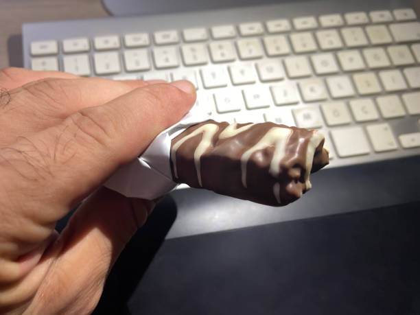 A hand holding open package chocolate bar in office. Eating chocolate while working